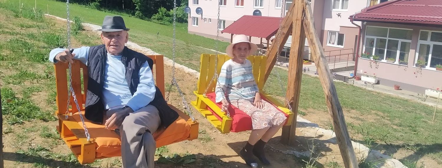 Dvoje stanara na ljuljaškama u dvorištu, iza njih zgrada doma sa cvijećem na prozorima